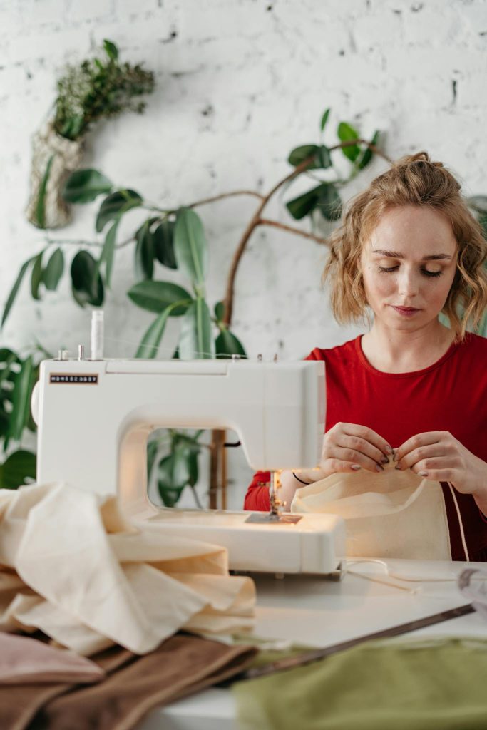 A woman in a red dress using a sewing machine indoors, surrounded by fabrics.